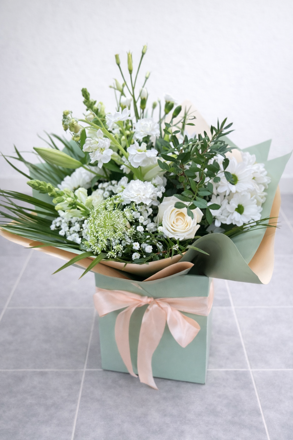 Bouquet of white and green flowers in a green box with a pink ribbon on a light grey background